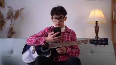 Attractive man playing on guitar in the bright room. Guy student try to learning the guitar lesson from the internet with his smartphone. Copy space