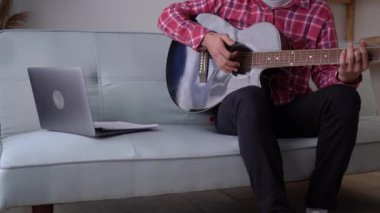 Handsome young man holding guitar and using his laptop in modern room. close up of male music coach or tutor play guitar have online video lesson on laptop at home. copy space
