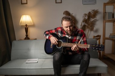 Handsome bearded man playing guitar while sitting on sofa in living room in the evening. Copy space