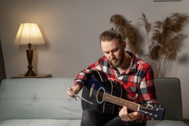 Composer with guitar examining sheets with notes sitting on sofa at home in the evening. Copy space