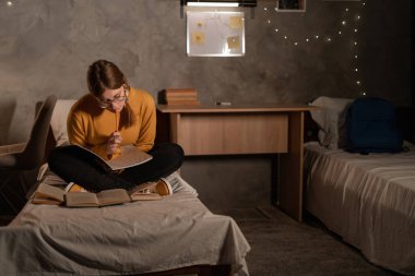 Student studying in the dormitory room sitting on the bed in the evening. Copy space
