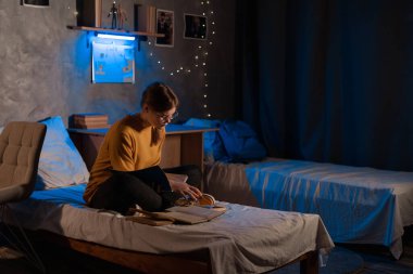 Young girl student doing the project looking for information in copybooks at dormitory room sitting on the bed at night. Copy space