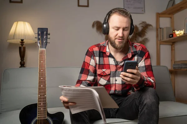 Happy man in headphones with music notes watching acoustic guitar tutorial on smart phone at home in the evening. Copy space