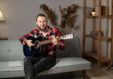 Smiling young man playing guitar while sitting on sofa late evening looking at camera. Copy space