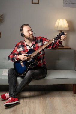 Young man playing guitar while sitting on sofa in living room in the evening. Copy space