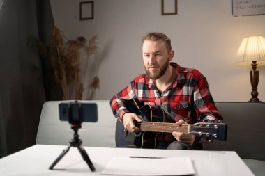 Student learning to play guitar at home or online tutor. Smiling man looks at guitar and makes video lesson for followers in living room. Copy space