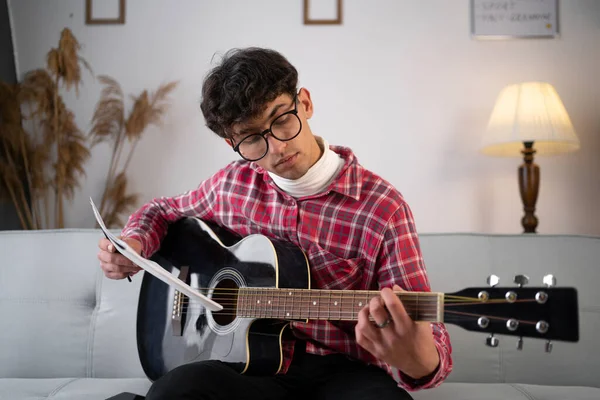 Male musician learning guitar lessons. Young guitarist holding a guitar and examining music sheets. Man studying music at home. Copy space