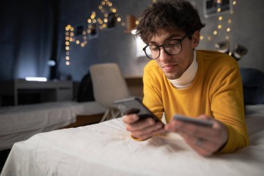 Portrait of happy 20s aged Arab man in casual clothing holding a credit card and mobile phone lying on bed at home in the evening. Finance, e-commerce, money banking online concept. Copy space