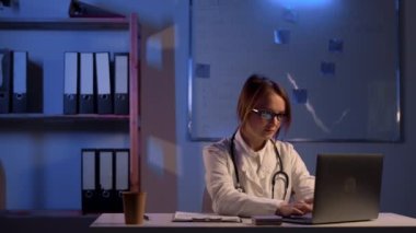 Female doctor in white coat and glasses typing on laptop computer at hospital during night shift. Healthcare, technology people and medicine concept