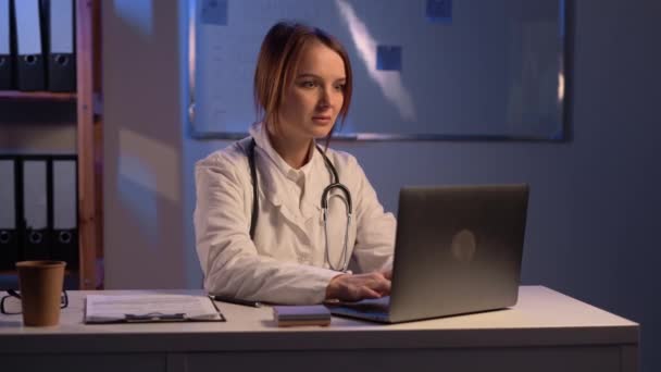 迷人的年轻医生穿着白衣坐在桌旁 上夜班时使用笔记本电脑 复制空间 — 图库视频影像