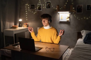 Calm teen man meditating with closed eyes in front of laptop pc at dormitory. Young male freelancer feeling peaceful and balanced, doing yoga sitting at his desk. Copy space