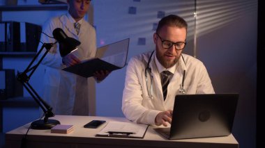 Young physician is talking to a millennial doctor in a medical health clinic while working at night shift. Colleagues discuss work on laptop in hospital office. Doctor and assistant. Copy space