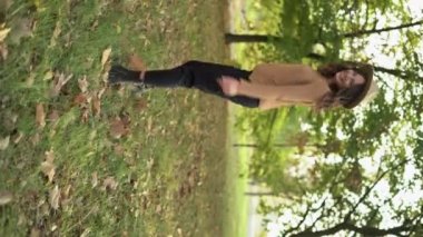 Happy girl looking behind while walking in autumn park. Beautiful woman relaxing at the park wearing sweater. Young smiling woman looking at camera. Vertical video