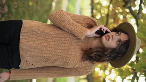 黄色の葉の背景に丸い帽子をかぶった携帯電話を呼び出しながら 公園内の秋の肖像画の女性 垂直ビデオ — ストック動画
