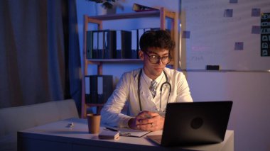 Steteskoplu beyaz önlüklü genç bir doktor, bilgisayarlı bilgisayarlı bilgisayar kullanarak sağlık kliniğindeki ya da hastanedeki hastalarla görüntülü konferans görüşmesi yapıyor, gece vardiyasında çalışıyor. Danışmanlık ve terapi.