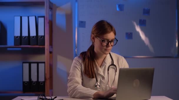 有魅力的医生穿着白衣坐在桌旁 在上夜班时使用笔记本电脑 复制空间 — 图库视频影像