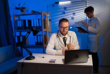 Male doctor working using laptop computer with colleague standing in background during night shift. Copy space
