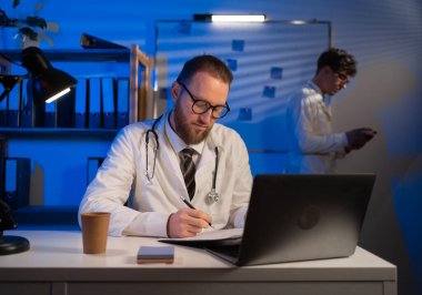 Adult doctor working night shift in hospital using laptop computer and his young therapist colleague standing in the background. Copy space