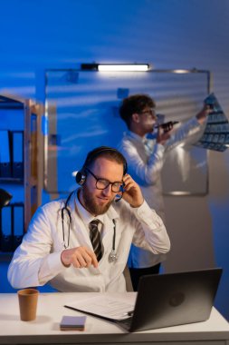 Doctor having video call with patient while his colleague looking at x ray scan in hand for medical diagnosis, working late at night. Radiography exam results for analysis and examination. Copy space