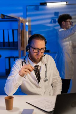 Doctor with headset consulting patient online at desk in clinic at night shift, his colleague young physician standing on background. Close up