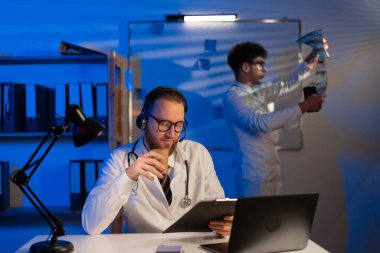 Male doctor working with document during night shift while his colleague looking at X-ray in medical office. Copy space