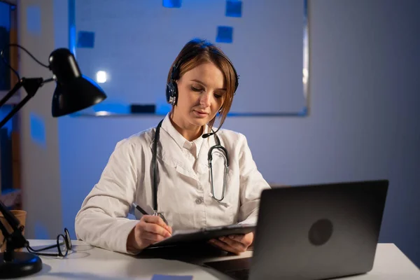 Doctor using headset with microphone working on laptop consulting patient online, therapist making video call at night, telemedicine concept. Copy space