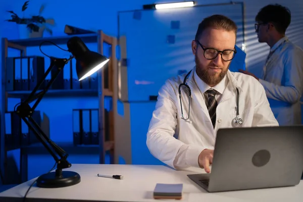 Young physician working with millennial doctor in a medical health clinic. Colleagues work on laptop computer in hospital office at night. Assistant and Medical Doctor. Copy space