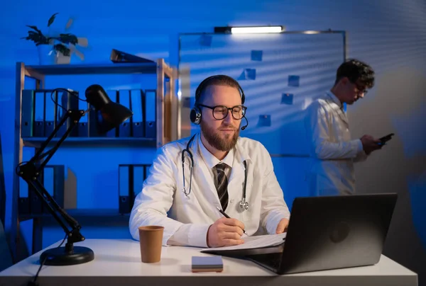 Male doctor wearing headset while using computer at desk in clinic with his colleague on background working at night shift. Copy space