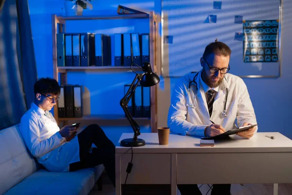 Young and adult male doctors in the hospital at night working with documents. Concentrated doctor over working sitting in a desk in a consultation. Copy space