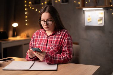 Young female student sitting at the table using smartphone when studying in dormitory late evening. Copy space