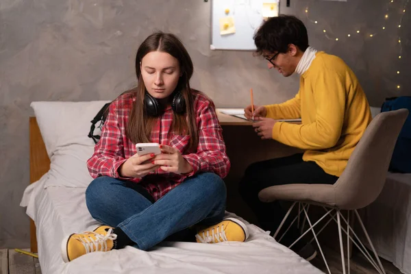 Serious students studying in dormitory room, girl watching online webinar using smartphone while sitting on bed and her boyfriend writing notes preparing for college exams. Copy space