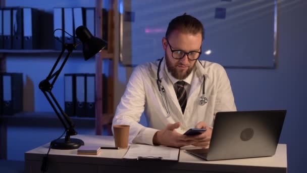 夜間の病院内のノートパソコンで動作するモバイルスマートフォンを使用して男性医師 電子健康記録システムEhr 遠隔医療の概念 — ストック動画