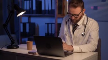 Serious male doctor using laptop and writing notes in medical journal sitting at desk. Medic physician with stethoscope working on computer at workplace at night shift. Copy space