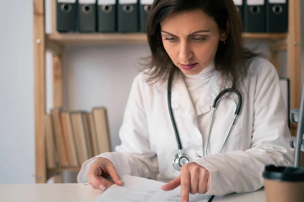 Concentrated female doctor reading document at table in clinic. Doctor cardiologist doing paperwork checking medical documents at workplace. Close up