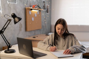 Young Asian girl student write on the notebook studying language online watch and listen to lecturer, webinar via video call e-learning at home. Copy space