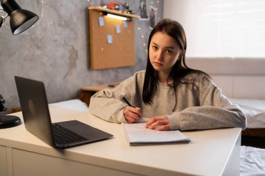 Portrait of a tired student learning online with laptop taking notes in a notebook sitting at her desk at home, looking at camera. mixed race asian chinese girl in dormitory. Copy space