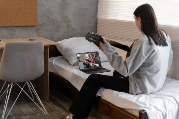 Focused girl playing acoustic guitar and watching online course on laptop while practicing at college dormitory. Online training, online classes. Copy space