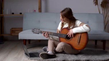 Online studying. Cute young woman is watching video tutorial, video classes how to play guitar, she sits on floor with acoustic guitar, using laptop to stream record music, chatting with friends.