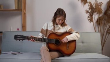 Young beautiful woman sitting on the couch and tuning a guitar strings in her music studio at home. Copy space