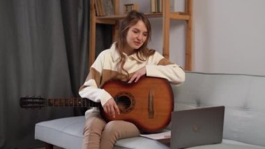 Young woman with guitar talking on webcam of laptop, learn to play with an online course, teacher online of guitar playing, sitting on the sofa at home. Copy space
