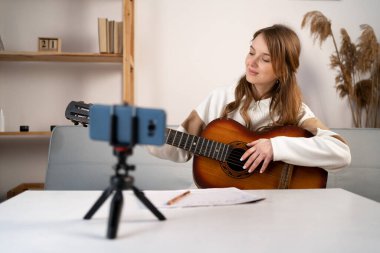 Music college student practicing acoustic guitar exercise, recording video on phone on tripod. Woman taking an online musical courses at home. Copy space