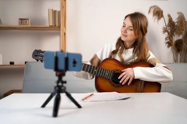 Music college student practicing acoustic guitar exercise, recording video on phone on tripod. Woman taking an online musical courses at home. Copy space