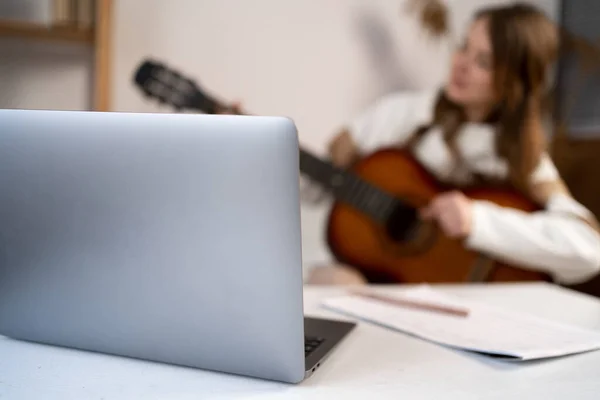 Music college female student practicing acoustic guitar exercise, reading notes from laptop computer, blurred. Woman taking an online music courses at home. Unfocused background, close up