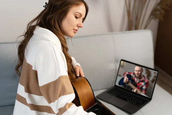 Focused woman playing acoustic guitar and watching online course on laptop while practicing at home. Concept of online training, online classes. Copy space