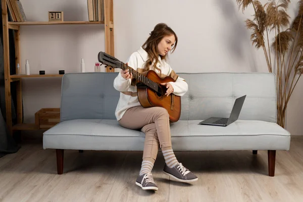 Woman practicing playing acoustic guitar in the living room, watching guitar tutorial on her laptop. Copy space
