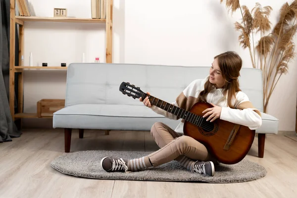 Photo of a young woman playing the guitar sitting in the living room. Girl singing a new song playing a stringed musical instrument. copy space