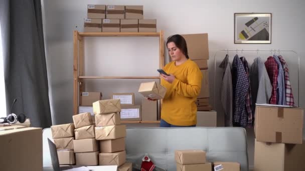 女性はスマートフォンで小包のバーコードをスキャンし 切手用の小さな段ボール箱を準備します 自宅のオフィスで働く中小企業のオーナー 中小企業コンセプト — ストック動画