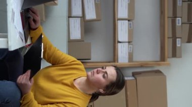 Genç işyeri sahibi kadın paket kutusu hazırlıyor ve dizüstü bilgisayardaki müşteriye teslimat için çevrimiçi ürün siparişlerini kontrol ediyor. İnternetten alışveriş konsepti. Dikey video