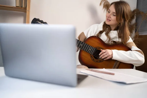 A young woman watches a video lesson on playing the guitar and practicing sitting on the sofa. Online training, online classes. Hobbies and leisure activities concept. Copy space