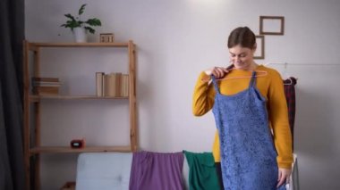 Beautiful attractive girl choosing clothes on clothes rack dressing looking herself in living room at house. Young woman think what to wear. Lifestyle women relax at home concept.